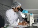 Scientist in a white lab coat and hairnet looking through a microscope at a laboratory bench indoors.