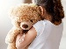 A young child seen from behind hugging a soft brown teddy bear tightly against their shoulder in gentle, natural light.