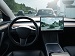 Interior of a modern car showing steering wheel and a central touchscreen with a camera view of traffic on a motorway, suggesting autonomous or driver-assist operation.