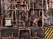 A dense network of old industrial metal pipes, valves and a large fan set against a worn machine wall.