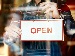 A shopkeeper in an apron holds an “Open” sign on a glass door, representing small businesses receiving new AI-powered support to stay efficient and welcoming.