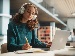 A teenage girl wearing headphones sits at a desk in a modern library, writing in a notebook while using a laptop, focused and studying with books and pens nearby.